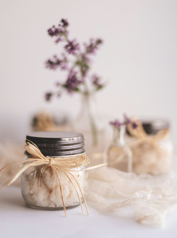 Pot en verre de sel de bain noué d'un ruban de raphia, entouré de fleurs violettes séchées dans un décor naturel épuré.