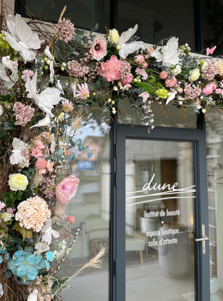 Porte de l'institut Dune encadrée d'un arc fleuri printanier aux tons roses, blancs et verts, avec papillons décoratifs.